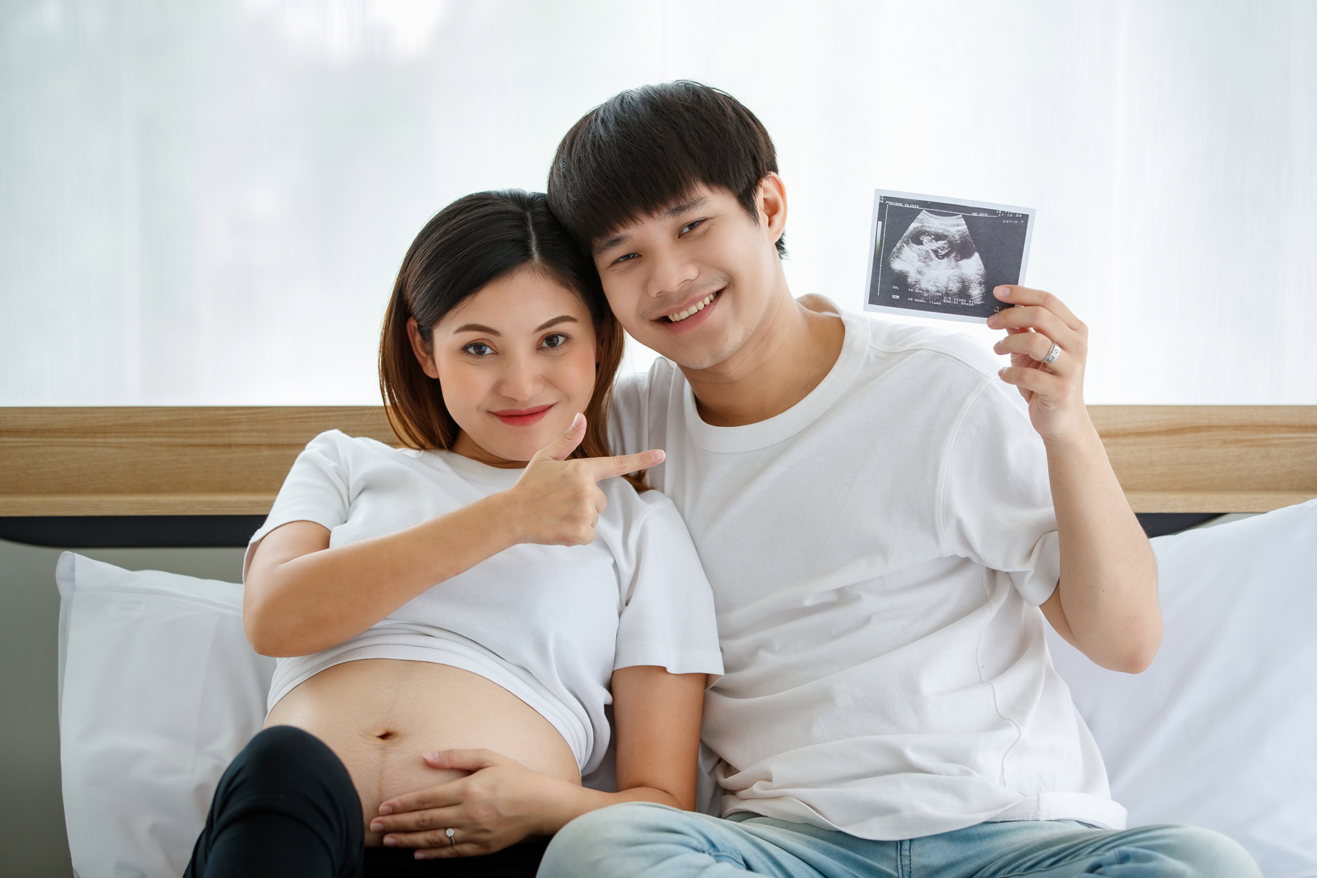 happiness young asian couple wearing casual dress smiling sitting bed together happy husband puts his arm around his pregnant wife showing ultrasound scan photo fetus